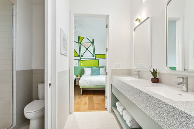 Bright modern en-suite bathroom with terrazzo double-sink vanity, potted succulent and white tile, toilet and glass shower to the left, open door revealing a bedroom with a bold green geometric accent wall and white bed on hardwood floors.