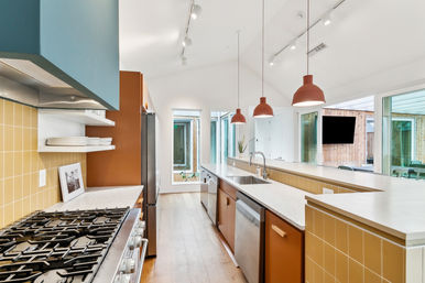 Bright open-concept modern kitchen with long island and breakfast bar, quartz countertops, stainless-steel appliances, mustard-yellow tile backsplash, terracotta pendant lights, hardwood floors and sliding glass doors to a courtyard