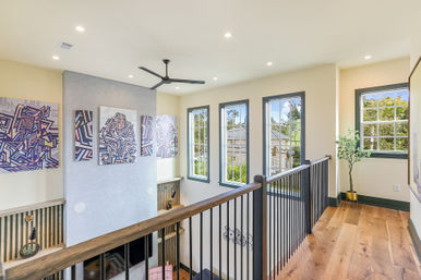 Airy sunlit upstairs landing in a contemporary home with hardwood floors, black metal railing, tall windows, abstract geometric gallery wall, ceiling fan and potted plant.