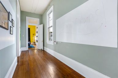 Sunlit hallway with polished hardwood floors, sage-green walls, a long whiteboard scrawled with 'New Orleans!', framed art and an open door revealing a bright yellow room.