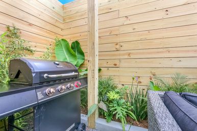 Backyard patio with black gas grill, wicker outdoor sofa, lush tropical plants and a modern horizontal wooden privacy fence.