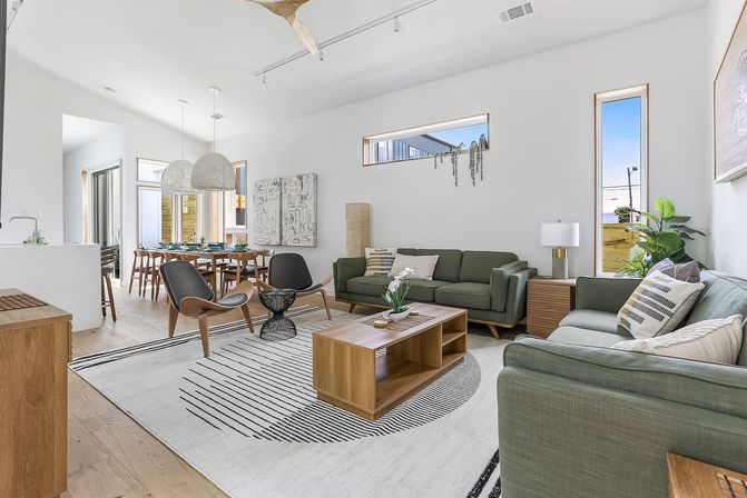 Sunlit open-plan living and dining room with mid-century modern green sofas, wooden coffee table, black-and-white geometric rug, sculptural lounge chairs, pendant lights over a set dining table, hardwood floors, tall narrow windows, and potted plants.