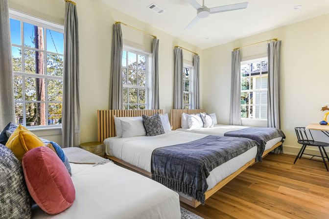 Sunlit modern bedroom with three beds dressed in white linens and gray throws, colorful accent pillows, tall multi-pane windows with gray curtains, ceiling fan, hardwood floors and a small desk — airy, rental-ready space.