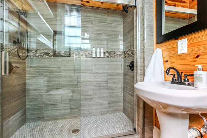Modern rustic bathroom with glass-enclosed walk-in shower, gray horizontal tile and mosaic accent strip, hexagon tile floor, matte black fixtures, and a white pedestal sink against wood-paneled wall.