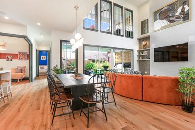 Bright modern open-plan dining and living room with wide-plank hardwood floors, long black dining table with metal chairs, orange curved sectional, globe pendant light, tall windows and sliding glass doors to a lush backyard patio.