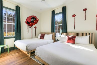 Sunlit modern guest bedroom with two platform beds in crisp white linens, slatted wooden headboards, emerald curtains, red accent pillows and bold red textile wall art, hardwood floor and windows showing palm trees.