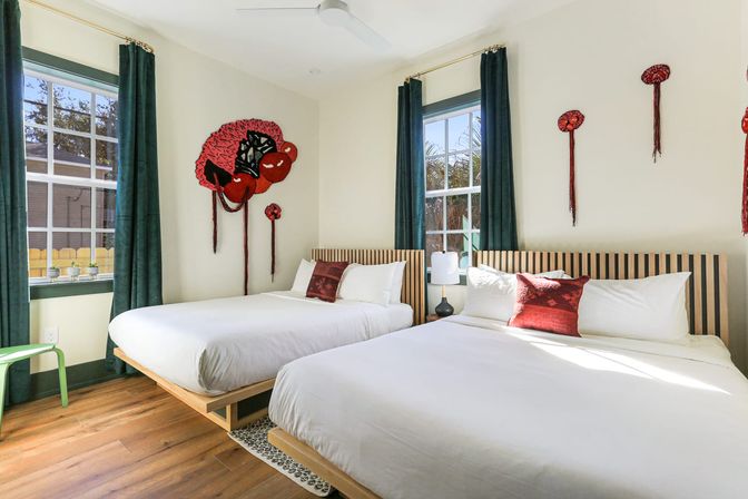 Sunlit modern guest bedroom with two platform beds in crisp white linens, slatted wooden headboards, emerald curtains, red accent pillows and bold red textile wall art, hardwood floor and windows showing palm trees.
