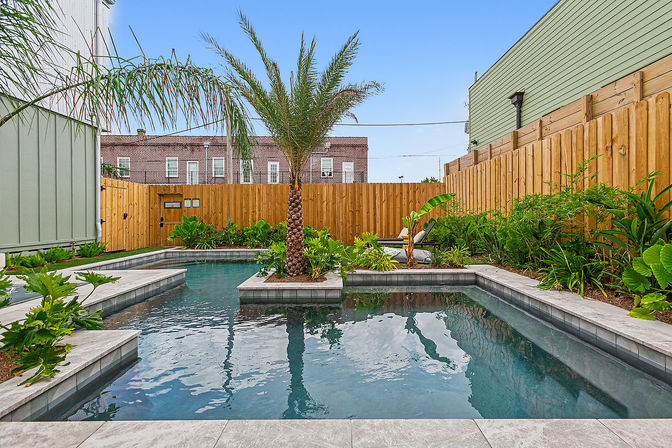 Urban backyard pool with a central palm island, gray stone decking, tropical plants, wooden privacy fence and brick rowhouses beyond