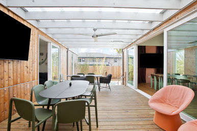 Bright covered wood deck patio with modern outdoor dining table and green chairs, sliding glass doors, coral lounge chair, ceiling fan, and fenced suburban backyard view.