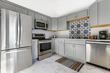 Bright modern kitchen with gray shaker cabinets, stainless steel French-door refrigerator, range, microwave and dishwasher, decorative blue-and-white patterned tile backsplash, white subway tile, marble-look floor and a striped woven rug.