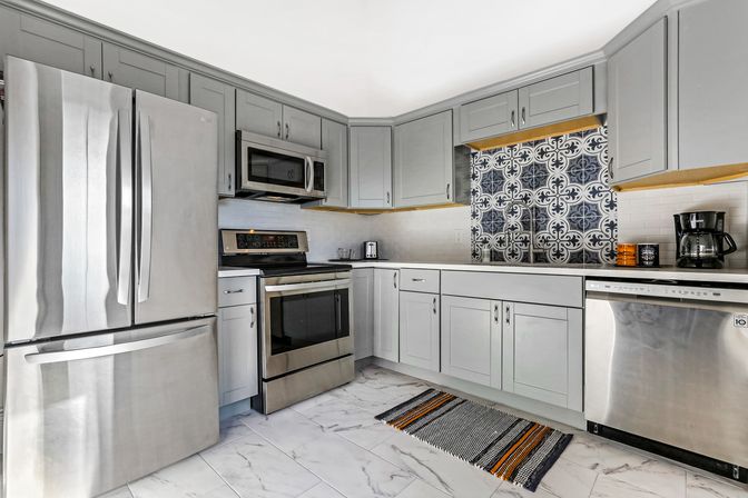 Bright modern kitchen with gray shaker cabinets, stainless steel French-door refrigerator, range, microwave and dishwasher, decorative blue-and-white patterned tile backsplash, white subway tile, marble-look floor and a striped woven rug.