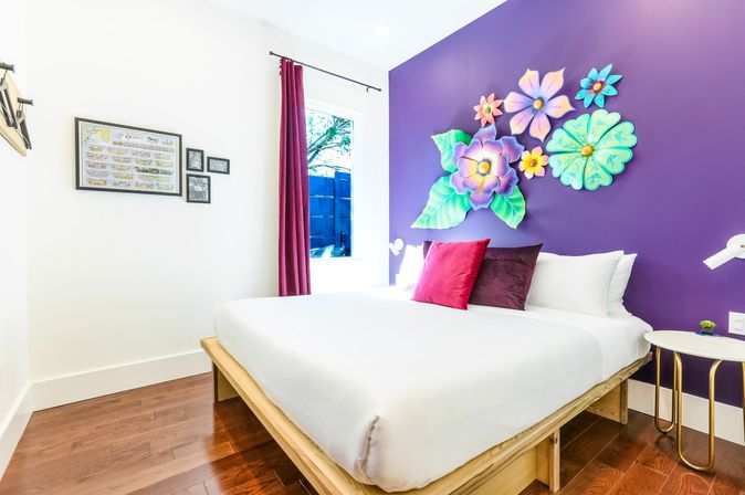 Sunlit modern bedroom with white bedding on a wooden platform bed, vibrant purple accent wall with colorful 3D floral wall art, red and plum throw pillows, hardwood floor, window with magenta curtain and a small round bedside table.