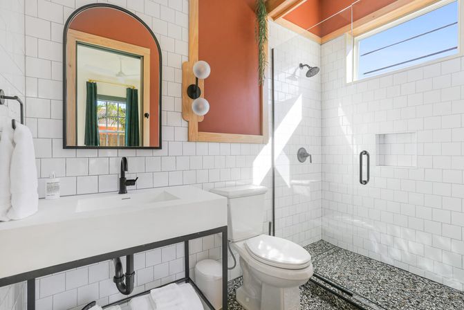 Sunlit modern bathroom with white subway tile, terrazzo shower floor, glass walk-in shower, matte black fixtures, arched mirror and warm rust-colored accent wall.