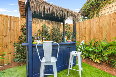 Backyard tiki-style bar with blue wooden counter and thatched roof, two white metal bar stools on green turf, bottles and tropical drinks on the bar, surrounded by a wooden privacy fence and lush tropical plants