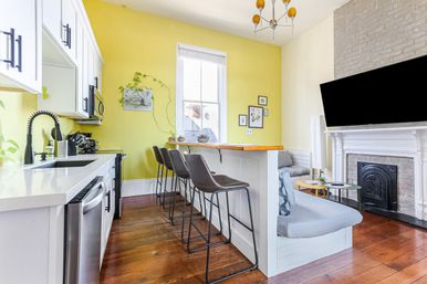 Cheerful open-plan kitchen and living area with a yellow accent wall, white cabinets and stainless appliances, wooden breakfast bar with four brown bar stools, hardwood floors, potted plants, and a white fireplace with mounted TV.