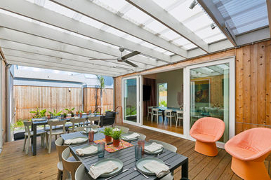 Sunlit covered backyard patio with wooden deck and translucent pergola, long black outdoor dining table set for a meal with plates and glasses, potted plants along a timber fence, sliding glass doors to the house, ceiling fan and two coral lounge chairs — modern outdoor dining and entertaining space.