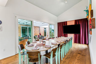 Spacious modern dining room with a long wooden communal table set for a meal, mint-green and wood chairs, sliding glass doors to a patio, vaulted white ceiling, hardwood floors, burgundy curtains and a wall-mounted TV — inviting gathering space.