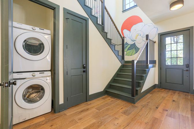 Bright mudroom entry with stacked washer and dryer in closet, green-painted staircase with large white floral mural, gray paneled door and oak hardwood floors.