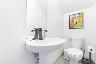 Bright modern white powder room interior with pedestal sink, matte-black waterfall faucet, toilet, colorful abstract wall art and gray stone-look floor, compact guest bathroom.