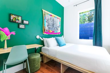 Cheerful modern guest bedroom with emerald green accent wall, single bed with crisp white linens and teal pillow, light wood platform bed and desk, mint chair and green velvet ottoman, colorful wall art and a window showing tropical greenery.