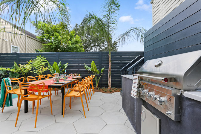 Modern backyard patio with long outdoor dining table set for a meal, bright orange chairs, built-in stainless steel grill, palm trees and dark horizontal privacy fence