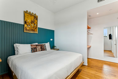 Bright modern bedroom with white king bed, teal vertical-paneled headboard, geometric accent pillow and yellow trumpet artwork above, hardwood floors and open closet with full-length mirror.