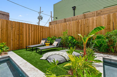 Sunny urban backyard oasis with a small plunge pool, two black lounge chairs on artificial turf, tropical plants and a tall wooden privacy fence.