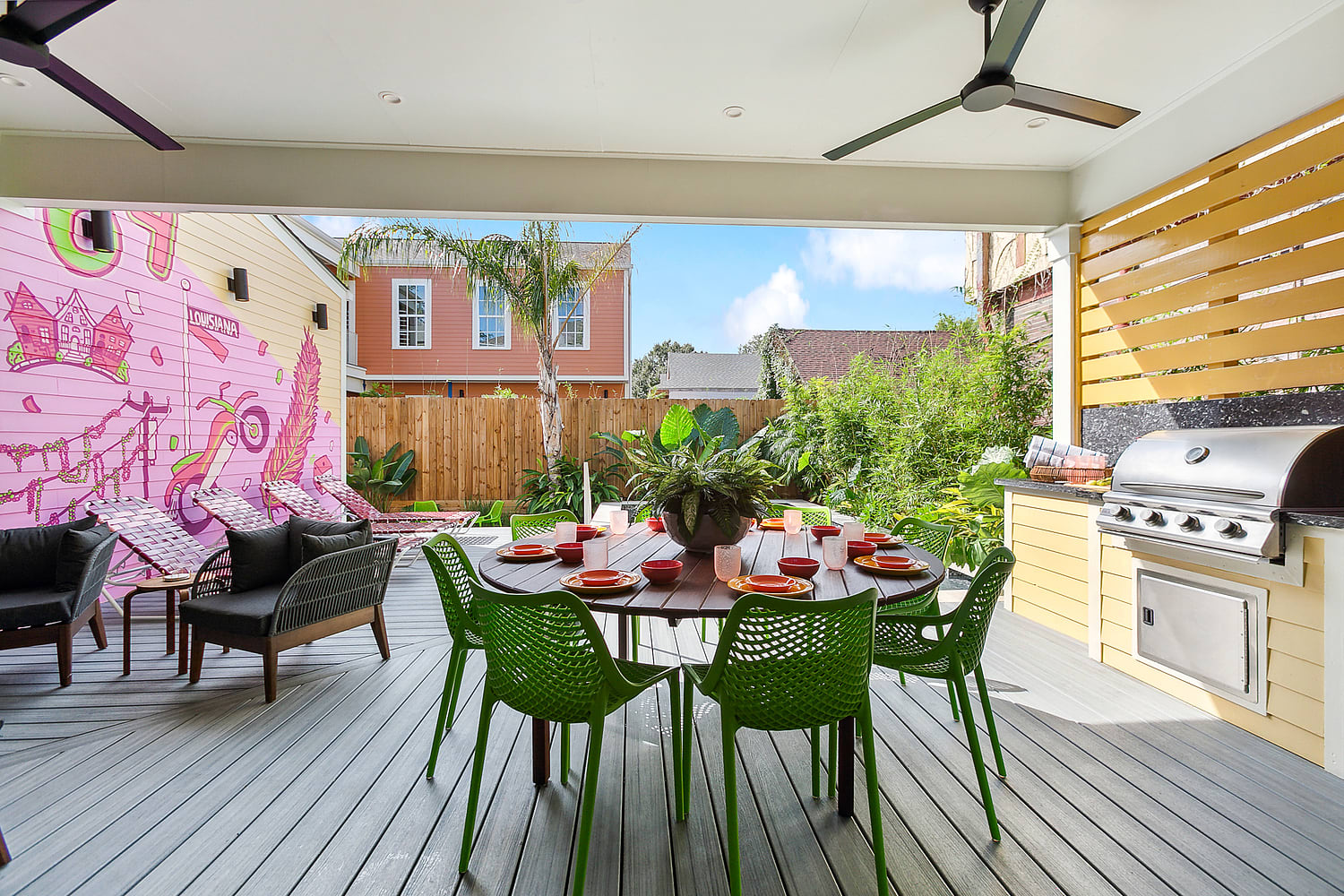 Covered backyard patio with round wooden dining table set for six with bright green chairs, stainless steel built-in grill, lounge seating, pink mural on the side wall and tropical plants and palm tree beyond a wooden fence