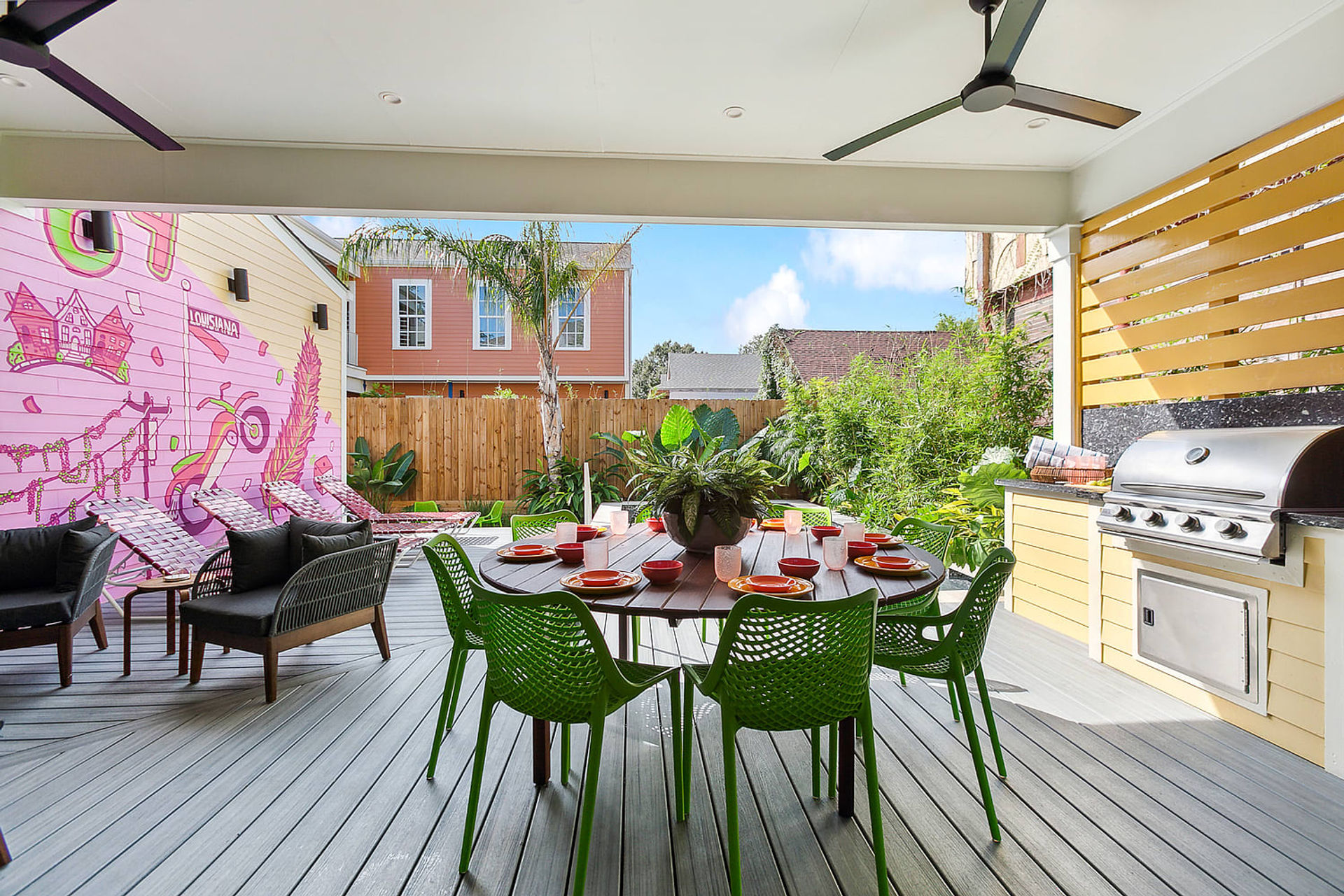 Covered backyard patio with round wooden dining table set for six with bright green chairs, stainless steel built-in grill, lounge seating, pink mural on the side wall and tropical plants and palm tree beyond a wooden fence