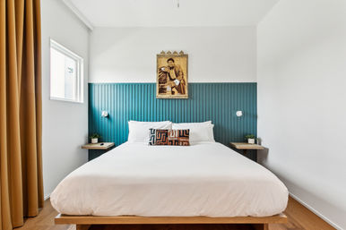 Bright modern bedroom with teal vertical-panel headboard, white platform king bed with geometric accent pillow, wood nightstands, mustard curtains, potted plants, and framed vintage portrait above the bed.