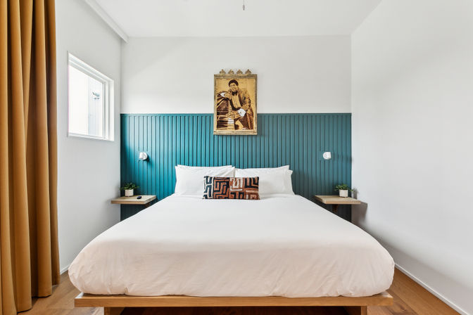 Bright modern bedroom with teal vertical-panel headboard, white platform king bed with geometric accent pillow, wood nightstands, mustard curtains, potted plants, and framed vintage portrait above the bed.