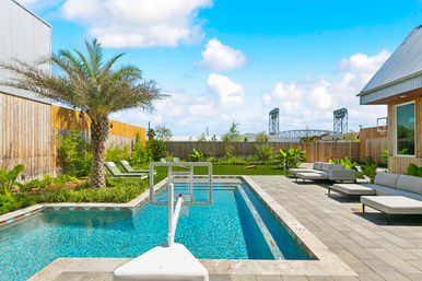 Sunny backyard pool with crystal-blue water and built-in basketball hoop, palm tree, lounge chairs, modern patio seating, grassy fenced yard and industrial lift bridge on the horizon.