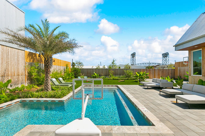 Sunny backyard pool with crystal-blue water and built-in basketball hoop, palm tree, lounge chairs, modern patio seating, grassy fenced yard and industrial lift bridge on the horizon.