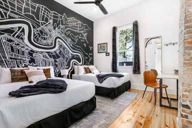 Modern boutique guest room with two double beds, striking black-and-white city map wall mural, hardwood floors, exposed brick column, desk with leather chair, window with curtains and ceiling fan — bright cozy vacation rental interior.