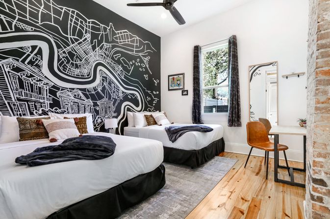 Modern boutique guest room with two double beds, striking black-and-white city map wall mural, hardwood floors, exposed brick column, desk with leather chair, window with curtains and ceiling fan — bright cozy vacation rental interior.