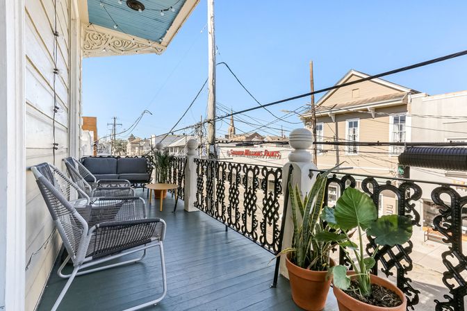 Sunny urban balcony with decorative wrought-iron railing, gray wicker chairs and sofa, small wood table, potted tropical plants and string lights under an ornate eave, overlooking a downtown street with low-rise buildings and power lines.