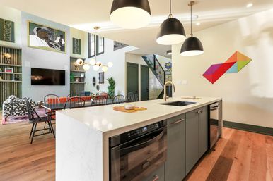 Bright open-concept kitchen and two-story living area with white marble island, gray cabinets, stainless appliances, black dome pendant lights, long dining table, hardwood floors and colorful geometric wall art.