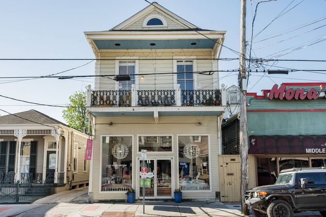 Charming sunlit two-story wooden building with a glass boutique storefront, ornate iron balcony above, neighboring shops, overhead utility lines and a parked SUV on the street.