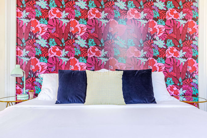 Bright bedroom interior with vibrant tropical floral wallpaper, neatly made bed with navy velvet and pale yellow pillows, white linens and gold side tables