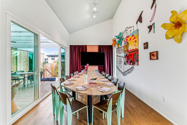Bright modern dining room with a long wooden table set for a meal, mint-accented chairs, colorful wall art and TV, and sliding glass doors to a backyard deck