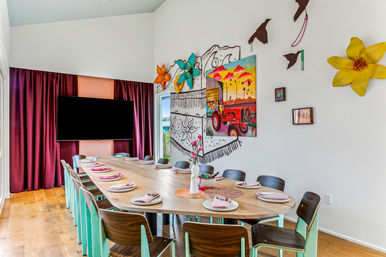 Bright modern dining room with a long oval wooden table set for twelve, mint-green chairs, colorful tractor mural and oversized flower wall art, wall-mounted TV, and plum curtains on wood floors
