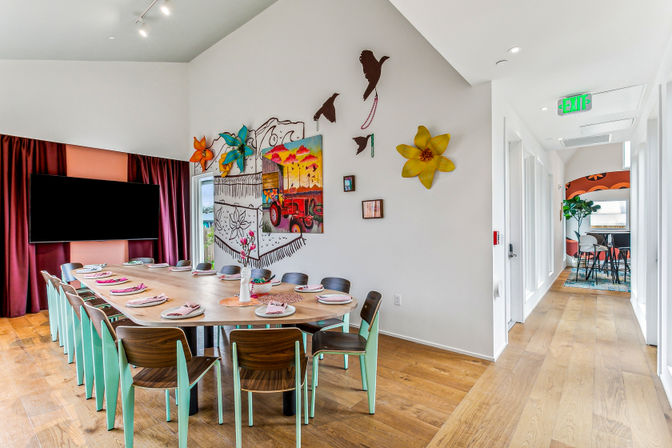 Bright modern clubhouse dining room with a long wooden table set for a meal, mint-green chairs, colorful wall murals and decor, large TV, hardwood floors and a sunlit hallway.