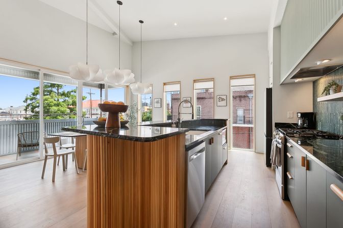 Bright modern open-plan kitchen in an urban apartment featuring a wood-slatted island with black stone countertop, stainless appliances, cloud-like pendant lights, floor-to-ceiling windows and balcony with city and tree views.