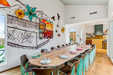 Bright open-plan dining room with a long wooden communal table set for 14, turquoise-legged chairs and colorful napkins, playful wall mural featuring oversized flowers and a tractor painting, hardwood floors and an open modern kitchen with yellow tile backsplash.