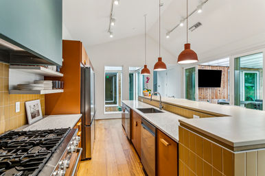 Bright open-concept modern kitchen with a long island and sink, three pendant lights, stainless steel range and fridge, speckled countertops, hardwood floors, and tall windows overlooking a courtyard.