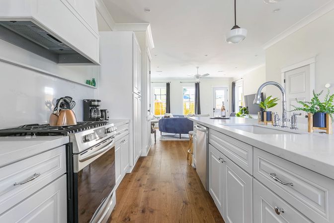 Bright modern white kitchen with quartz island and sink, stainless steel range and appliances, warm hardwood floors, and open-concept living room beyond
