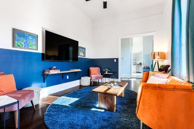 Sunlit modern living room with a navy blue half-wall, wall-mounted TV, round deep-blue rug, wooden coffee table, orange velvet sofa, pink accent chairs and playful pops of color leading to an open kitchen.