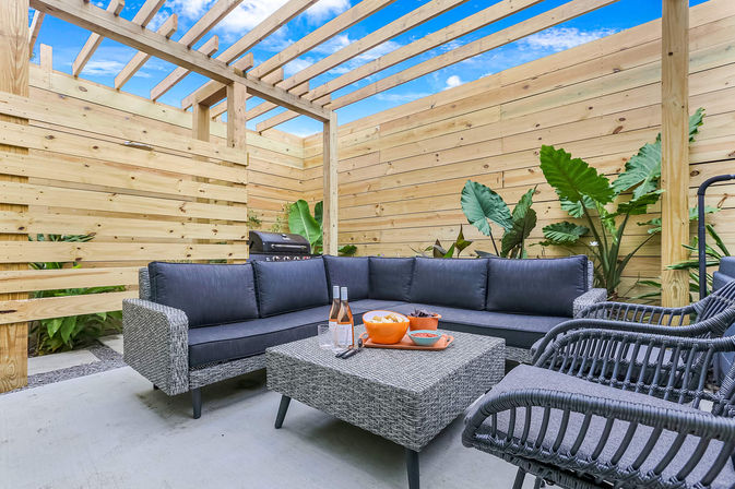 Cozy modern backyard patio with gray wicker sectional and chairs under a wooden pergola, coffee table with snacks and drinks, and tropical plants.