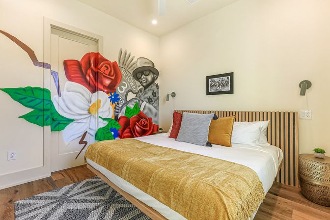 Bright modern bedroom with a large bed, mustard throw and slatted wood headboard; bold wall mural of red roses, a white flower and a grayscale figure in a hat and sunglasses; patterned rug and round wooden nightstand.
