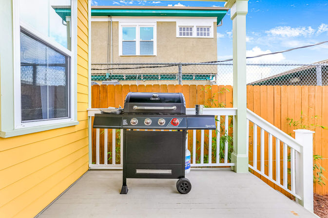 Sunny residential backyard deck with a black propane gas grill on a covered yellow house porch, white railings and wooden privacy fence with neighboring building backdrop